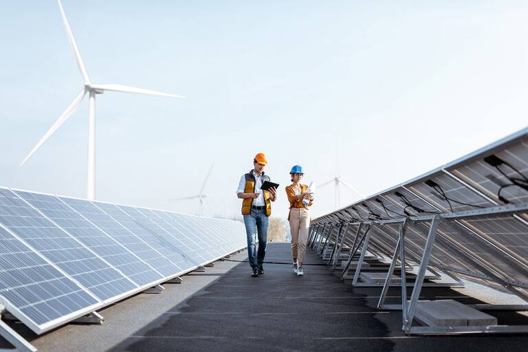 Zwei Personen mit Schutzhelmen inspizieren eine Solaranlage, während im Hintergrund Windkraftanlagen zu sehen sind.