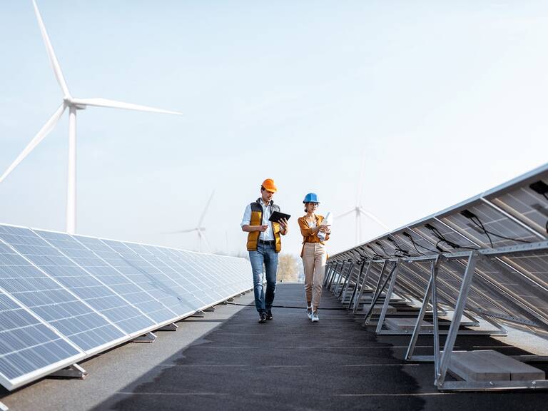 Zwei Personen mit Schutzhelmen inspizieren eine Solaranlage, während im Hintergrund Windkraftanlagen zu sehen sind.