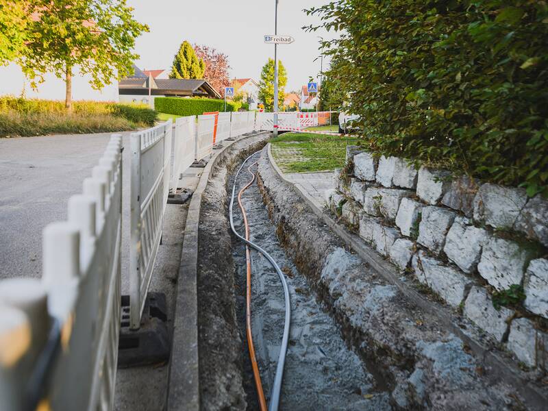 Ein offener Graben verläuft entlang einer Straße, die durch Absperrungen gesichert ist, während Glasfaserkabel verlegt werden.