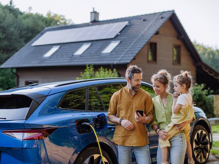 Eine dreiköpfige Familie steht vor einem blauen Elektroauto, das geladen wird, mit einem modernen Haus im Hintergrund.