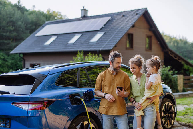 Eine dreiköpfige Familie steht vor einem blauen Elektroauto, das geladen wird, mit einem modernen Haus im Hintergrund.