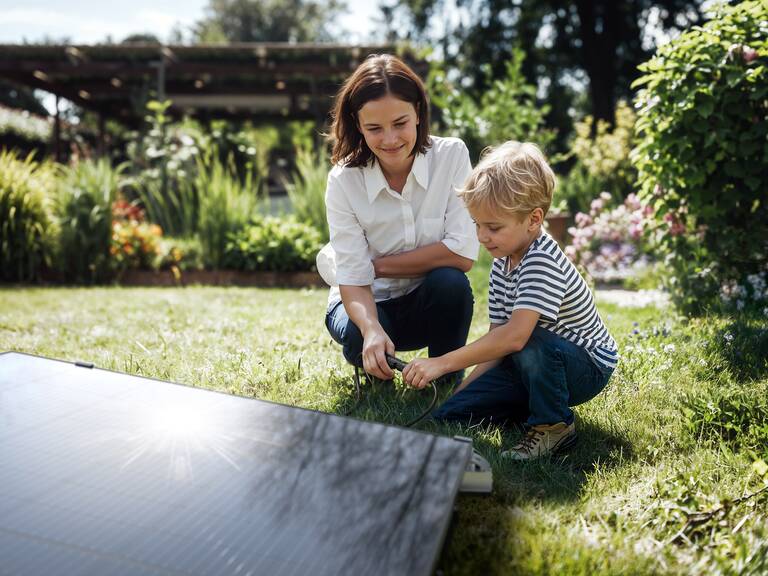 Eine Frau und ein Kind sitzen im Garten auf dem Gras neben einem Solarpanel und beschäftigen sich gemeinsam mit einem Gegenstand.