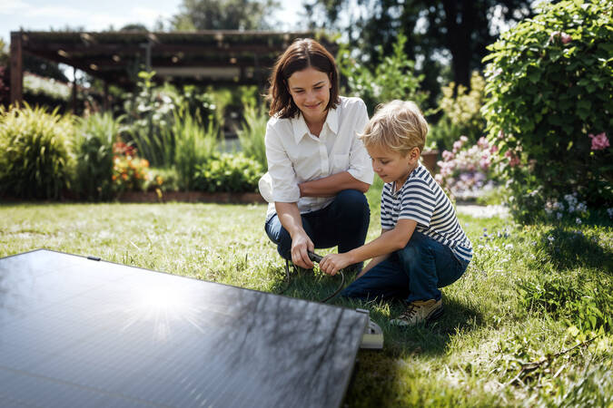 Eine Frau und ein Kind sitzen im Garten auf dem Gras neben einem Solarpanel und beschäftigen sich gemeinsam mit einem Gegenstand.