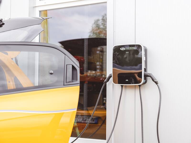 Ein gelbes Auto steht neben einer Ladestation an einer Gebäudewand, und das Ladekabel ist angeschlossen.