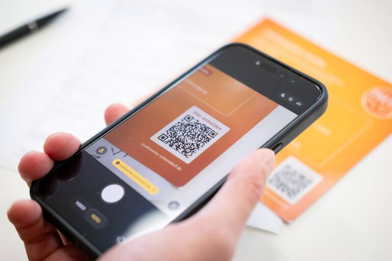 Eine Person scannt mit einem Smartphone einen QR-Code von einer orangefarbenen Broschüre.
