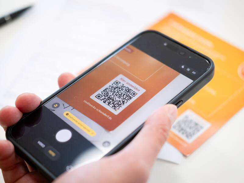 Eine Person scannt mit einem Smartphone einen QR-Code von einer orangefarbenen Broschüre.