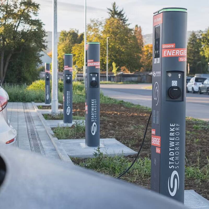 Eine Reihe von Elektroauto-Ladestationen auf einem Parkplatz bei Tageslicht, mit geparkten Autos im Hintergrund und Grünflächen daneben.