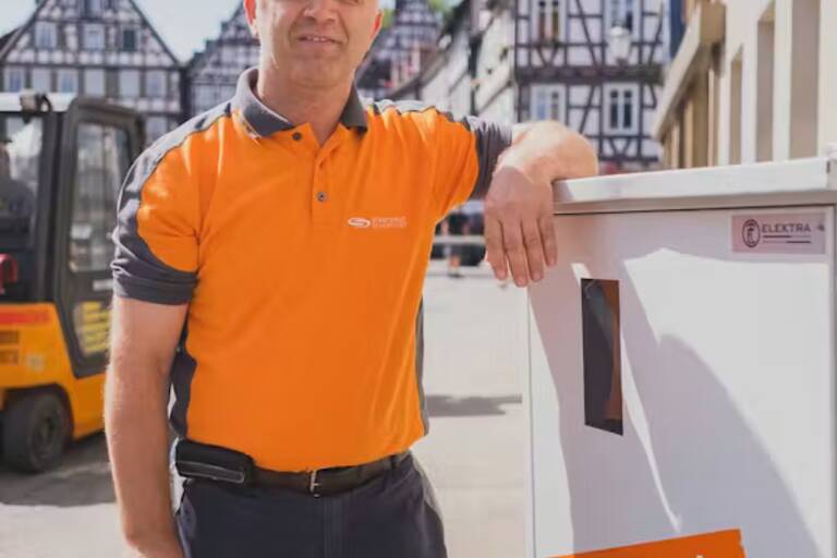 Ein Mann in einem orangefarbenen Polohemd steht neben einem Schaltkasten mit der Aufschrift "Die Energie für hier!" in einer Fachwerkhaus-umgebenen Straße.