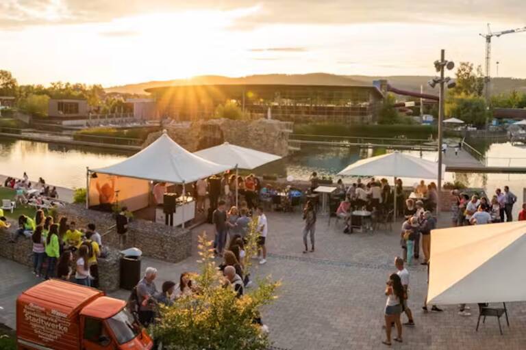 Menschen versammeln sich an einem sonnigen Abend auf einem Platz mit Zelten und einem Gewässer im Hintergrund für eine Veranstaltung.