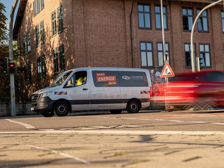 Ein Lieferwagen mit der Aufschrift "Unsere Energie für Sie" steht an einer Ampel vor einem Backsteingebäude, während ein rotes Auto vorbeifährt.