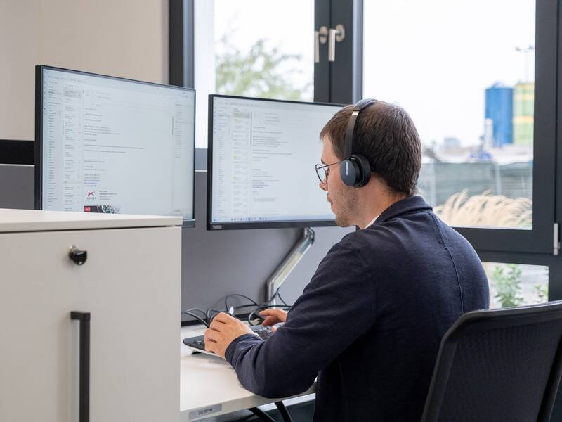 Eine Person sitzt mit Kopfhörern vor zwei Computerbildschirmen in einem Büro.