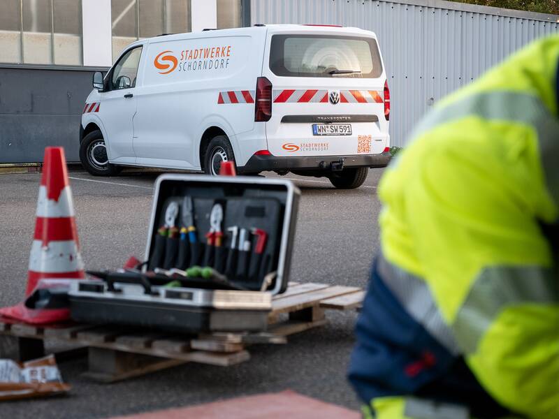 Ein weißes Transportfahrzeug der Stadtwerke Schorndorf steht auf einem Parkplatz, umgeben von Verkehrskegeln und einem Werkzeugkoffer im Vordergrund, während eine Person in Warnschutzkleidung arbeitet.