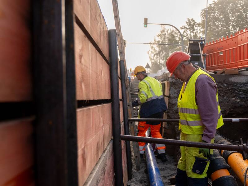 Zwei Bauarbeiter in Schutzkleidung arbeiten in einem tiefen Graben, umgeben von Baugerüsten und Rohren.