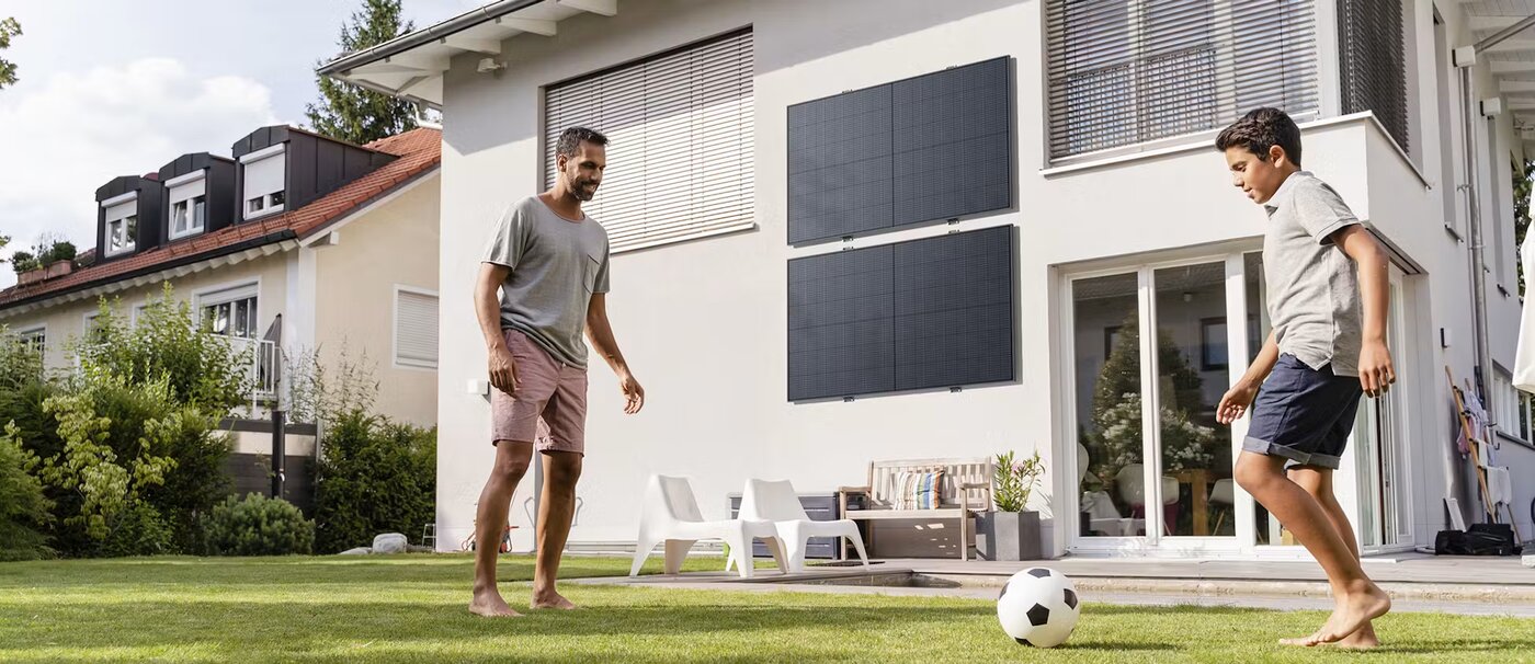 Ein Mann und ein Junge spielen mit einem Fußball im Garten vor einem Haus mit Solarmodulen an der Fassade.